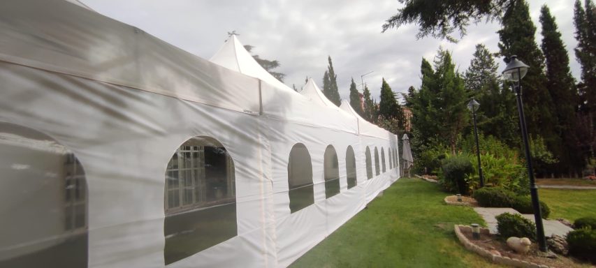 Carpa blanca con ventanas, ubicada en un área ajardinada con árboles y césped.